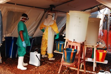 A hygienist steps in to the undressing tent to begin removing multiple items of protective clothing. The sprayer (left) guides the often exhausted colleague step by step through the undressing process. The protective layers can create stifling conditions that are difficult to withstand for too long. To avoid overheating and fatigue, staff working in the high risk zone must limit the time they spend in the isolation ward. ©Fathema Murtaza, MSF